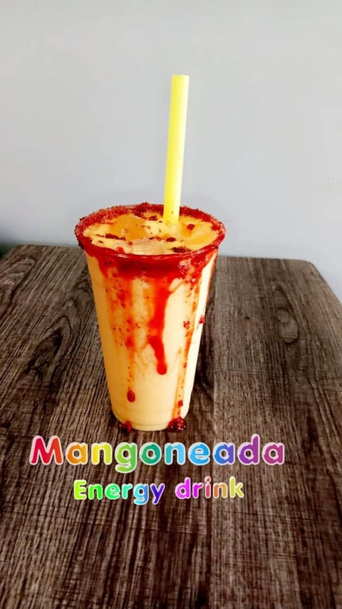 Mangoneada energy drink in a clear cup with red chili rim and yellow straw on wooden surface