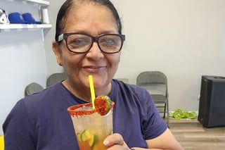 Woman wearing glasses and navy shirt smiling while holding a colorful cocktail with a yellow straw and rimmed glass