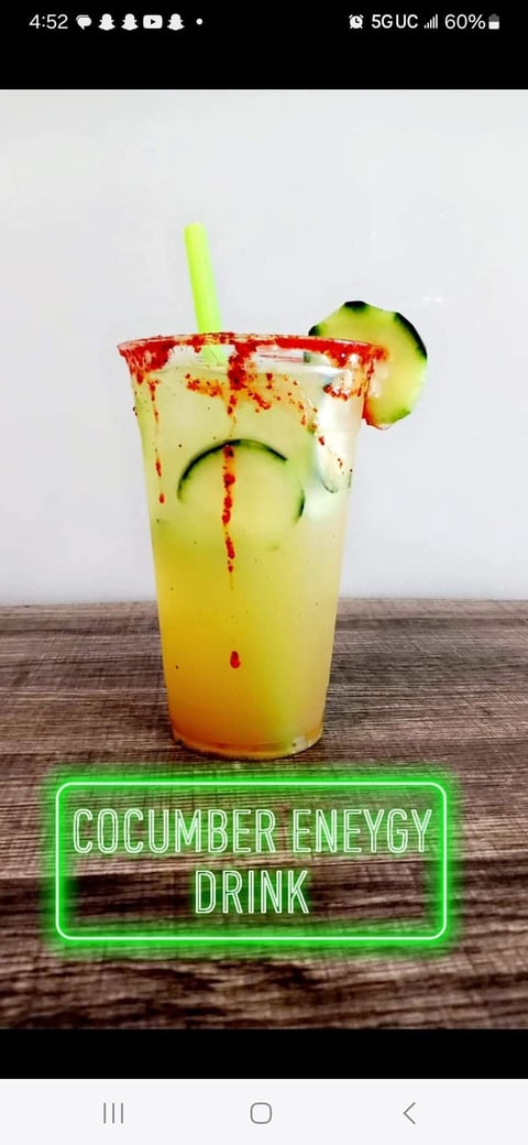 Cucumber energy drink in a clear glass with red spiced rim, cucumber slice garnish, and neon green text label on wooden surface