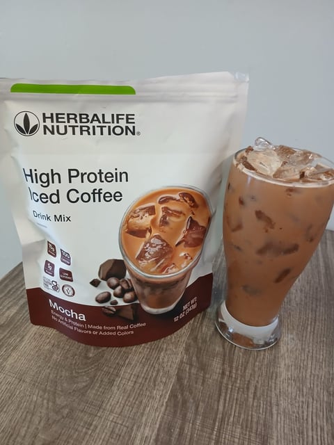 Herbalife High Protein Iced Coffee mocha drink mix package next to a glass of prepared iced coffee beverage on wooden surface