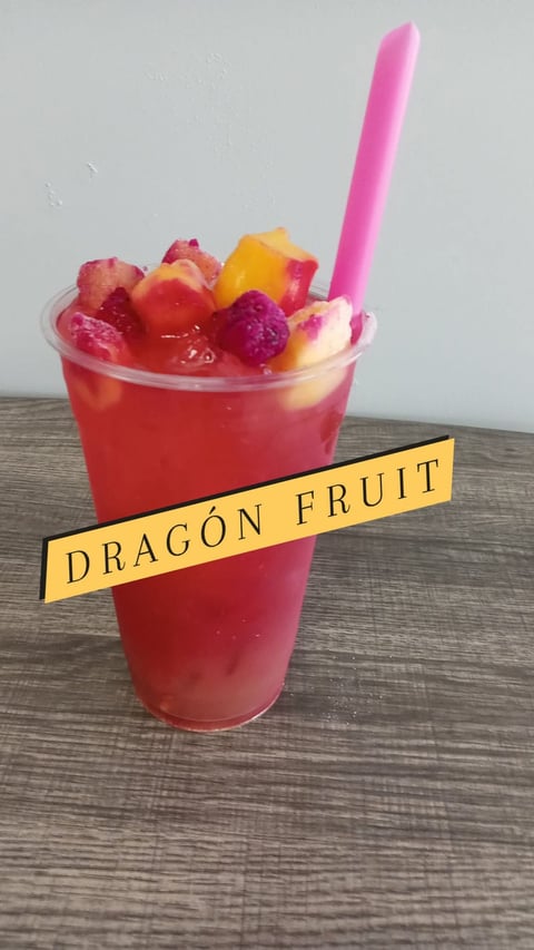 A vibrant pink dragon fruit beverage in a clear cup topped with dragon fruit pieces, a yellow label reading Dragon Fruit, and a pink straw.