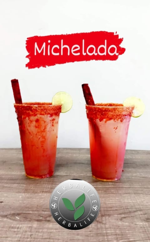Two red Michelada cocktails with lime slices and straws on wooden surface with Herbalife logo