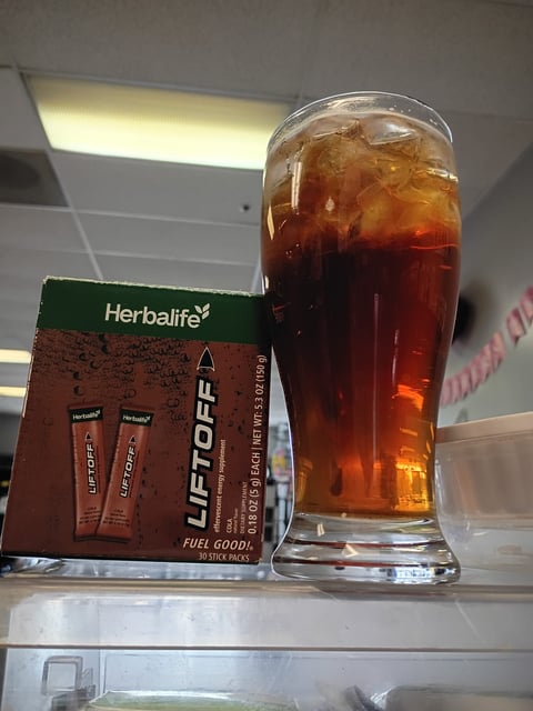 Herbalife LiftOff energy drink packets next to a tall glass of cola with ice on a shelf