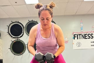 Woman in pink workout clothes holding dumbbells in a gym with fitness posters and exercise equipment on the walls