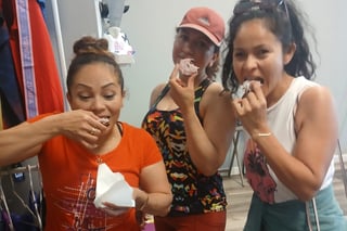 Three young women enjoying snacks together indoors, smiling and eating while standing in casual clothing.