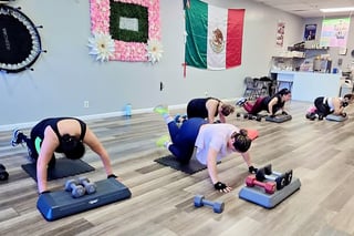 Group fitness class with participants doing push-up exercises on step platforms in a bright studio with decorative flowers and flags on walls