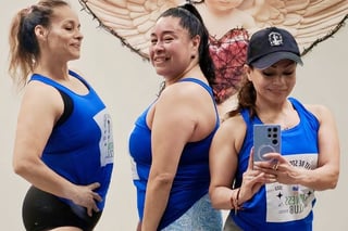 Three women in blue athletic tanks and race bibs smiling together indoors, with one holding a phone for a selfie